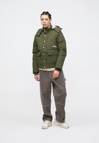 Casaco acolchoado verde-oliva com dois bolsos frontais, botões de pressão e gola alta, combinado com calças largas cinzentas e sapatilhas claras.