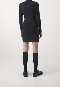 Vestido negro de canalé con mangas largas y cuello alto, combinado con calcetas negras hasta la rodilla y zapatos negros, sobre un fondo neutro.