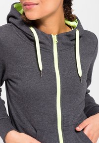 Sudadera con cremallera gris y forro interior verde neón, con cordón. Fabricada en un tejido suave, cuenta con dos bolsillos frontales y un collar alto.