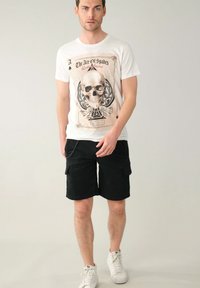 T-shirt bianca con una grande grafica di teschio decorata. Pantaloni cargo neri e sneakers bianche completano l'outfit.
