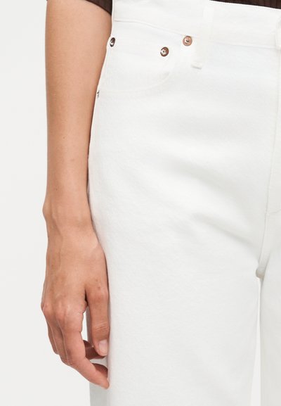 Αριστερό χέρι που ακουμπάει δίπλα σε λευκό τζιν ψηλής μέσης με μεταλλικά καρφιά και μπροστινή τσέπη, σε ένα ανοιχτό, ουδέτερο φόντο.