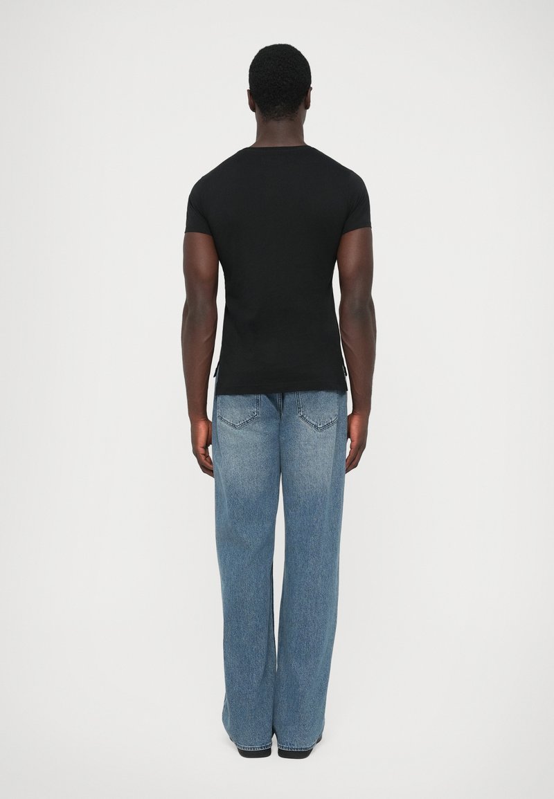 Uomo con pelle scura che indossa una maglietta nera aderente e jeans blu larghi, in piedi con le spalle rivolte verso di noi su uno sfondo bianco semplice.
