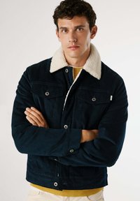 Giacca di corduroy blu marino con colletto in pelliccia shearling crema, dotata di due tasche sul petto e chiusura con bottoni, indossata sopra un maglione giallo.