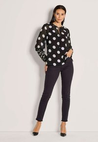 Frau trägt eine schwarze, durchsichtige Bluse mit Punkten und langen Ärmeln, schwarze enge Hosen und schwarze spitze Absätze und steht vor einer hellen Wand.