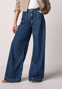 Weite Jeans in dunkelblauem Denim mit hoher Taille, vorderen Taschen und sichtbaren Nähten, kombiniert mit einer neutralen, kurzen Jacke.
