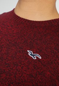 Punainen kuvioitu neulepaita pyöreällä pääntiellä. Rinnassa on valkoinen logo ääriviiva. Teksturoitu kangas, jossa on vähäistä kiiltoa.