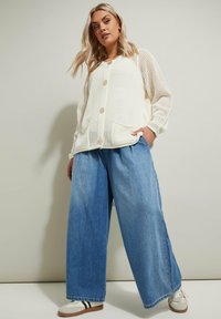 Cardigan leggero in maglia crema con grandi bottoni e tasche frontali, abbinato a jeans blu a gamba larga e sneakers bianche.