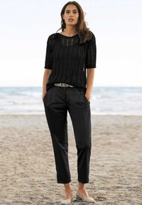 Zwart gebreid top met korte mouwen en een losse pasvorm, gecombineerd met zwarte tapered broek en lichtbeige ballerina's, staande op een zandstrand.