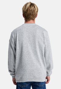 Sudadera gris claro con una tela texturizada, cuello redondo y puños de canalé. Presenta un pequeño parche con el logo en la manga izquierda.