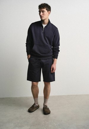 Junger Mann steht und trägt einen dunkelblauen Reißverschluss-Sweatshirt, passende Shorts, beige Socken und braune Slipper vor einem schlichten Hintergrund.