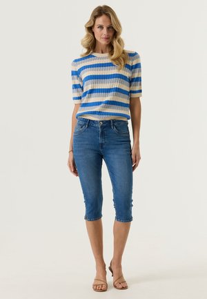 Frau steht und trägt einen blau-beigen, gestreiften Pullover mit kurzen Ärmeln, mittel-lange Jeans und beige Sandalen mit offenen Zehen vor einem einfarbigen Hintergrund.