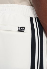 Balti džinsai su juodomis šoninėmis juostelėmis ir EA7 logotipu ant kišenės. Atrodo, kad medžiaga yra minkšta, o elastinga juosmens juosta užtikrina patogumą.