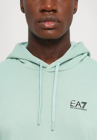 Mint green hoodie made of soft material, featuring a drawstring hood and embroidered EA7 logo on the chest. Smooth texture, casual design.