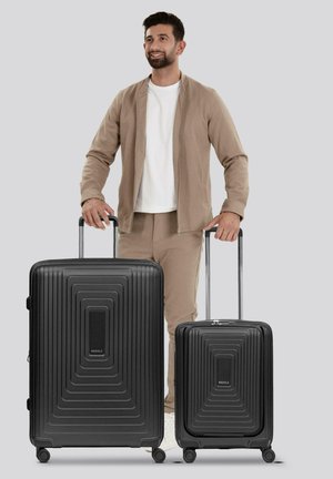Homme en tenue beige se tenant entre deux valises à coque rigide noires avec poignées extensibles sur fond gris.