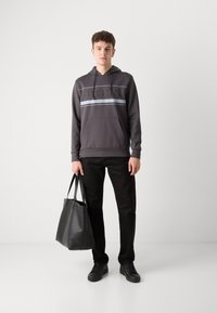Felpa grigia con cappuccio caratterizzata da un logo "BOSS" testurizzato, abbinata a pantaloni neri e una borsa a tracolla in pelle nera. Tessuto liscio con polsini a costine.