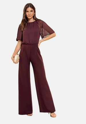 Donna con una tuta bordeaux con maniche, tiene una pochette beige, in piedi con una mano sui fianchi e indossa sandali dorati.
