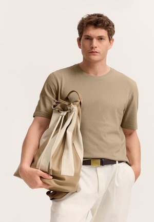 Junger Mann trägt beige strukturiertes Kurzarmhemd und weiße Hose, hält einen beigen und cremefarben gestreiften Rucksack mit Kordelzug.