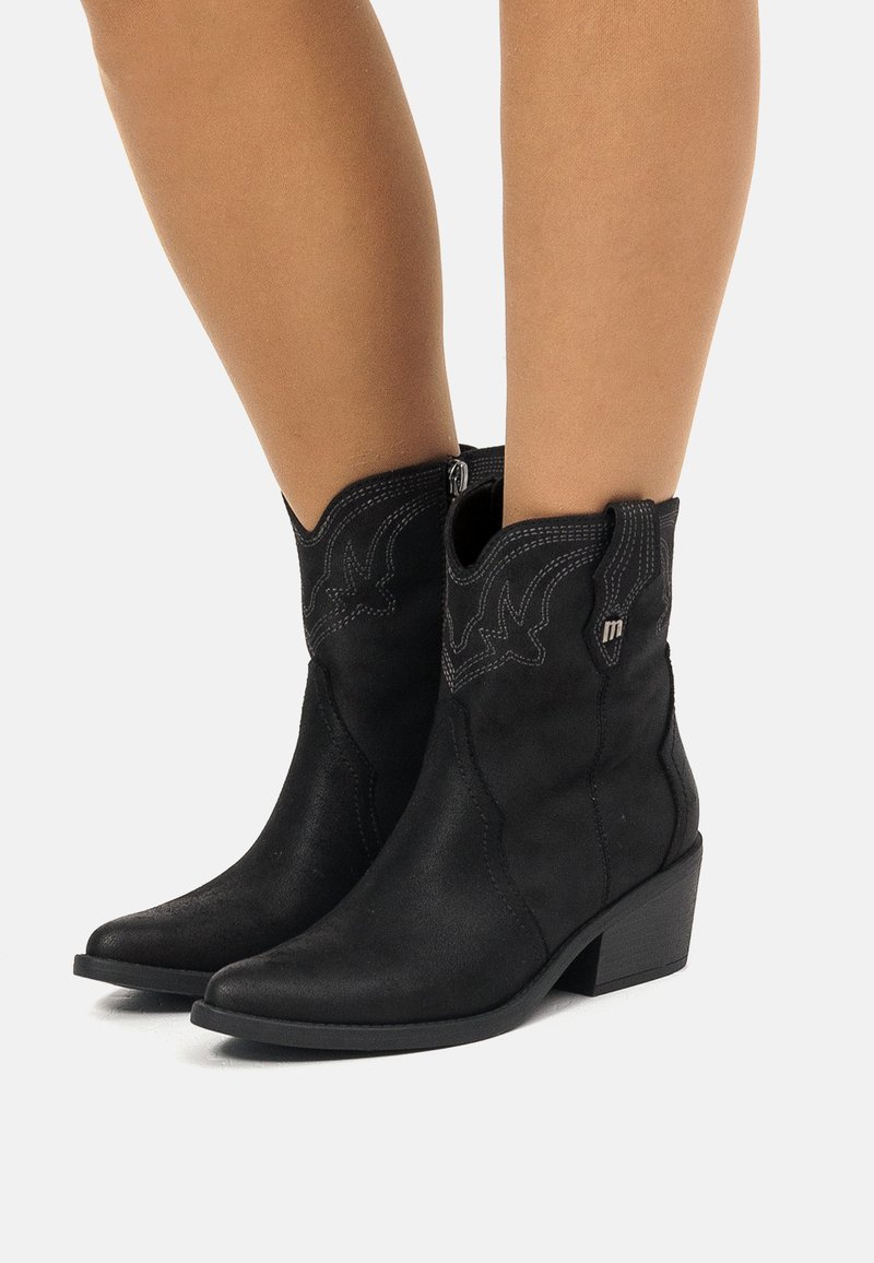Bottes de cowboy en suedine noires mi-mollet avec des coutures décoratives, des talons bas empilés et des zippers latéraux portées sur des jambes nues.