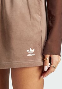 Brown cotton dress with a smooth texture, featuring an Adidas logo in white at the hem. The dress is paired with a sheer brown sleeve.