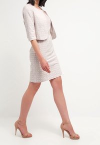 Robe rose clair ajustée avec motif floral texturé, associée à une veste courte assortie. Portée avec des sandales à talons hauts couleur nude.