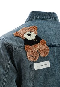 Chaqueta de mezclilla con un parche de oso de peluche texturizado en marrón y blanco, vistiendo una camiseta negra y con una etiqueta blanca en el lateral.