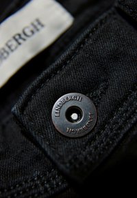 Tejido de mezclilla negro con un primer plano de un broche de metal que presenta "Lindbergh" grabado alrededor de un agujero central. Superficie texturizada y costuras visibles.