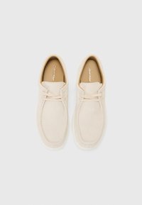 Scarpe slip-on in suede beige con dettagli cuciti e suola in gomma bianca, con una fodera interna marrone e un logo sulla soletta.