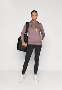 Fioletowa bluza z kapturem z złotym logo, czarne legginsy i białe trampki; do tego czarny plecak. Materiał jest miękki, elastyczny i sportowy.