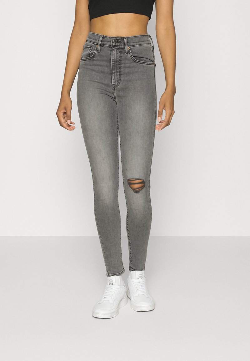 Jean denim gris taille haute avec un genou déchiré, coupe slim, et design classique à cinq poches, porté avec des baskets blanches.