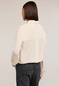 Blusa crema con maniche lunghe, tessuto morbido, colletto strutturato e pannello orizzontale sul retro, abbinata a jeans grigio scuro.
