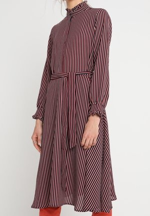 Frau trägt ein langärmliges, hochgeschlossenes Kleid mit vertikalen schwarzen, weißen und roten Streifen und einem gebundenen Gürtel an der Taille.
