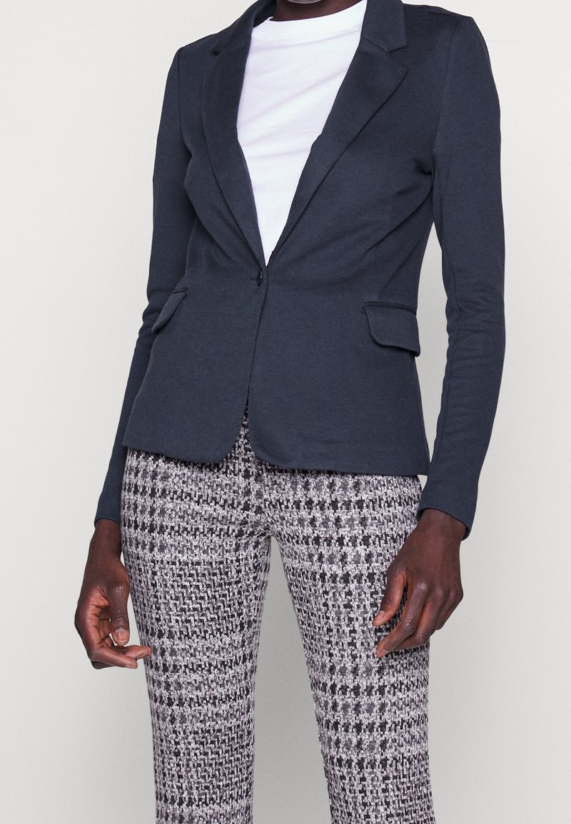 Blazer blu navy su misura con rever a lancia, due tasche frontali e una texture liscia, abbinato a pantaloni slim-fit con motivo nero e bianco.