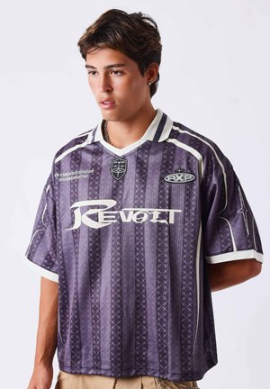 Junger Mann mit kurzen braunen Haaren, der ein lila gestreiftes Sporttrikot mit weißen Logos und Text trägt, vor einem einfarbigen hellen Hintergrund steht.