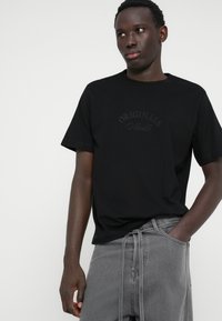 Camiseta negra de manga corta con texto en relieve "ORIGINALS". Material de algodón suave, corte holgado y cuello redondo. Combinada con jeans grises.