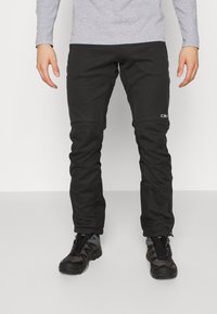 Schwarze Softshell-Hosen mit figurnaher Passform, ausgestattet mit Kniepolsterung und einem Logo am Oberschenkel. Kombiniert mit schwarzen Wanderschuhen.