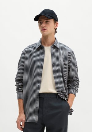 Młody mężczyzna w czarnej czapce, koszuli w pionowe paski zapinanej na guziki, na beżowym t-shircie oraz czarnych spodniach, z jedną ręką w kieszeni.