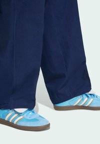 Light blue athletic shoes with white and gold stripes, paired with dark navy pants. Shoes feature mesh and suede materials with textured soles.