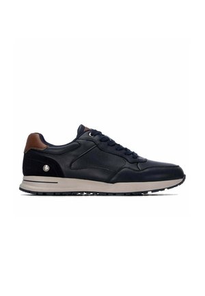 Zapatilla deportiva baja en negro y marrón con cordones negros, laterales texturizados, suela beige y un pequeño logotipo redondo cerca del talón, vista desde el exterior.