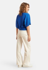 Blauer gestrickter Pullover mit überschnittenen Schultern, kombiniert mit weiten, cremefarbenen High-Waist-Hosen. Die Schuhe sind weiße Sneaker mit glatter Textur.
