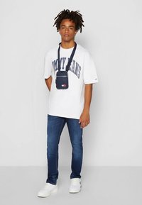 Balta oversize T-krekls ar uzrakstu „TOMMY JEANS”, tumši zili džinsi un baltas sporta kurpes. No siksnas karājas maza tumši zila plecu soma.