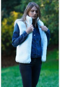Gilet en fausse fourrure blanche avec un col haut, superposé à une veste en denim foncé. Le mannequin porte un jean noir, affichant un look moderne et épuré.