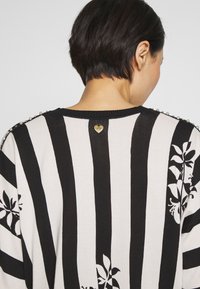 Personne portant un pull à rayures verticales noires et blanches avec des motifs floraux et des détails en perles sur les épaules, vue de dos.