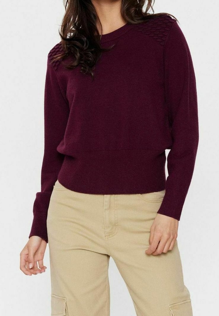 Kobieta w długim rękawie, w burgundowym swetrze prążkowanym i beżowych spodniach cargo, stojąca z rękami swobodnie przy bokach.