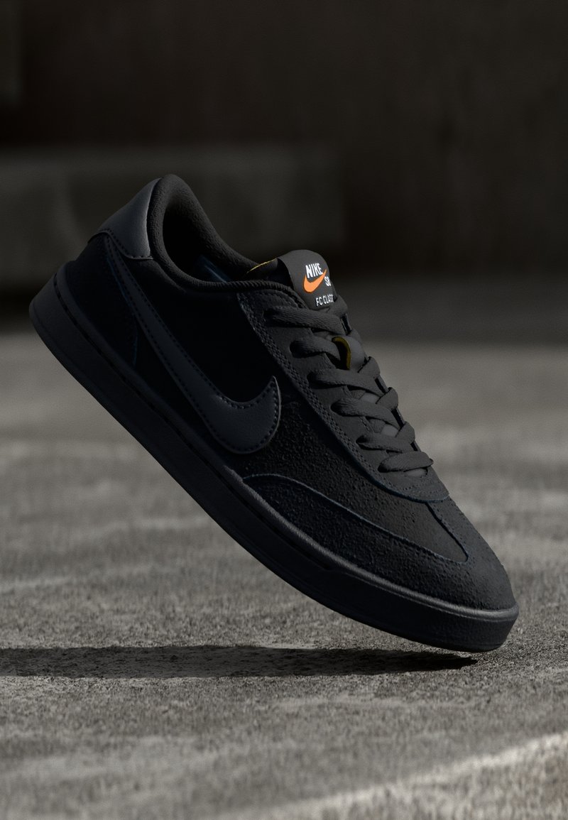 Baskets basses noires Nike en suède, avec un swoosh noir et un logo orange sur la languette, flottant au-dessus d'une surface grise en béton.