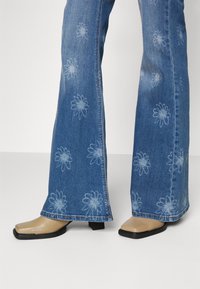 Blauwe flared jeans met lichte bloemenprints, gedragen over beige vierkante lage hakken op een effen witte achtergrond.