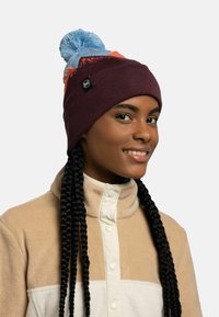 Gorro de punto en color burdeos con un pompón azul claro, que presenta un diseño colorido en la parte superior y una etiqueta de marca en el lateral.