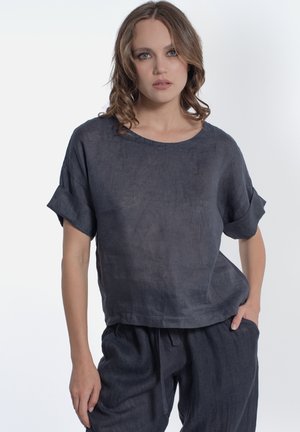 Donna con capelli ricci che indossa una maglia corta ampia grigio scuro e pantaloni con coulisse abbinati su sfondo bianco uniforme.