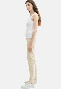 Biała bezrękawnik, beżowe spodnie, jasne sneakersy. Gładki materiał, dopasowany krój i casualowy design. Widok boczny na jednolitym tle.