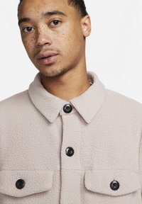 Lyserosa fleece-jakke med krage, frontknapper og to brystlommer. Har en myk tekstur og svarte knappeaccenter.