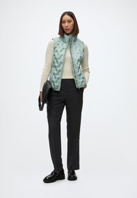 Svetlozelená prešívaná vesta, krémový rebrovaný sveter, tmavosivé nohavice, čierna kabelka a masívne čierne topánky. Hladká textúra a priliehavý strih.